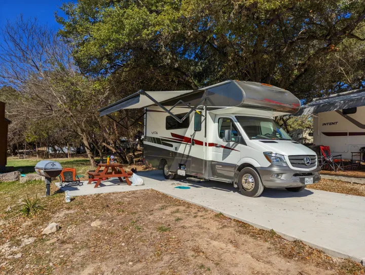 An RV rental with an extended awning is parked at a sunny campsite, complete with a picnic table and grill.. Winnebago Vita 2019