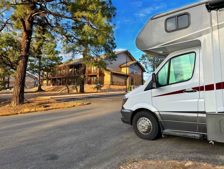 A white RV rental is parked on an asphalt road, with multi-story buildings and tall pine trees in the background under a blue sky.. Winnebago Vita 2019