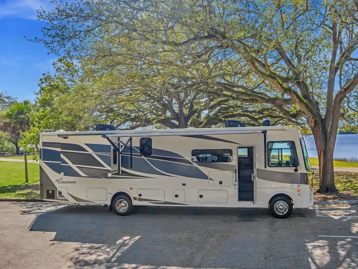 Spacious Class A rv rental parked under large trees in a scenic park by a lake, ideal for travel and leisure.. Coachmen Mirada 2021