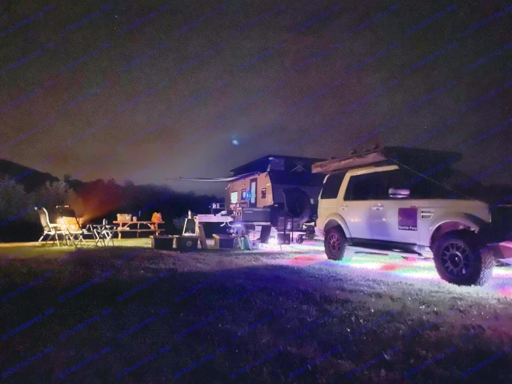 Nighttime camping with an RV rental setup. A brightly lit off-road camper and SUV stand on colorful ground lights, with chairs nearby.. Other Other 2022