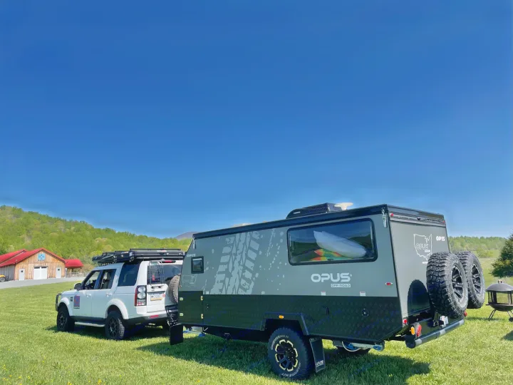 A white SUV tows a grey off-road camper trailer on a grassy field. Mountains & barn visible. Seek an RV rental for adventure!. Other Other 2022