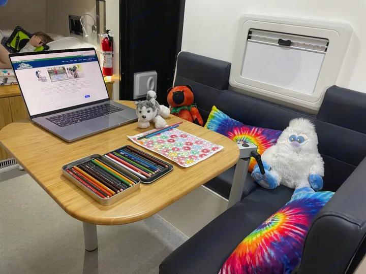 Inside a spacious RV rental: A laptop, coloring page, & plush toys fill a table, while a person naps in the bunk.. Other Other 2022