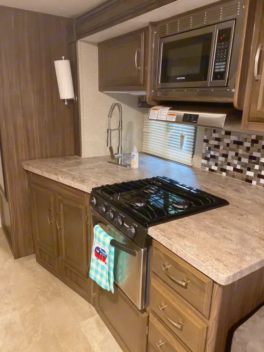 Functional RV rental kitchen with a microwave, gas stove, sink, wood-look cabinets, and mosaic tile backsplash.. Coachmen Other 2019