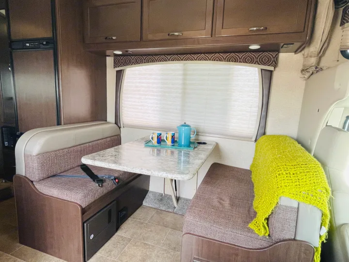 An RV rental interior showing a comfortable dinette with a table, benches, overhead cabinets, and a window with blinds.. Thor Chateau 2017