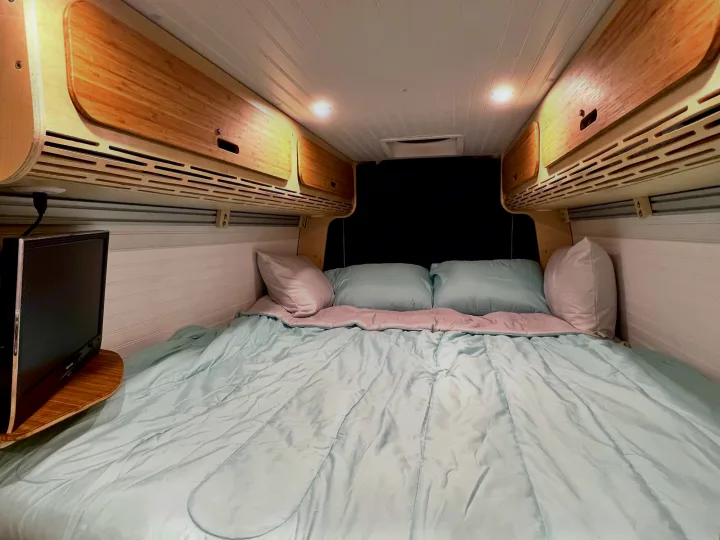 Cozy RV rental interior: A made bed with light blue comforter, overhead wood cabinets, TV, and white paneling.. Mercedes-benz Sprinter 2016