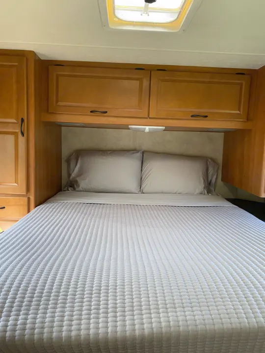 An RV rental's cozy bedroom, featuring a light grey bed with pillows, wooden overhead cabinets, and side storage.. Thor Majestic 2013