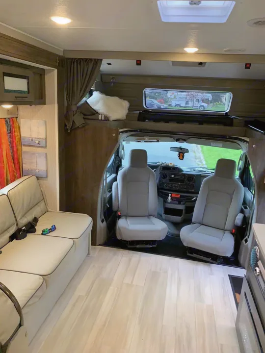 Inside a modern RV rental: light wood-look floor, comfortable sofa, overhead bed, and driver's cab visible.. Coachmen Leprechaun 2022