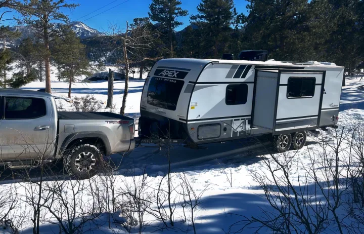 A silver truck tows an Apex Nano Off-Grid RV rental through a snowy mountain landscape, ready for winter adventure.. Forest River Coachmen Apex Nano Off-Grid 2022