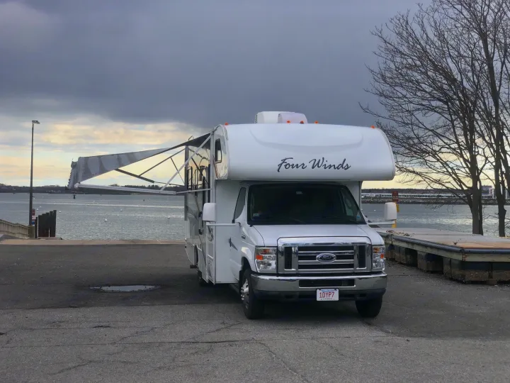 White "Four Winds" RV rental with extended awning, parked by a dock next to the water under a cloudy sky.. Four Winds Four Winds Motorhome 2011