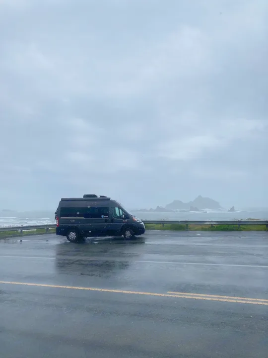 Dark RV rental parked on a wet coastal road, overlooking a stormy ocean with distant rocky islets under a grey sky.. Thor Motor Coach Rize 2022