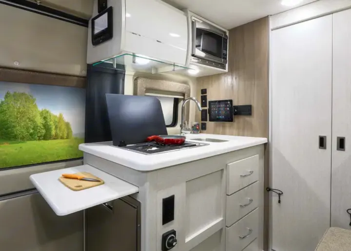 RV rental's compact kitchen: cooktop, sink, microwave, pull-out counter, and window view of nature.. Thor Motor Coach Rize 2022
