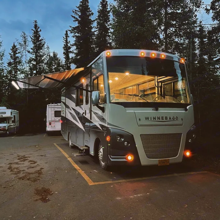 Brightly lit Winnebago RV rental parked for the night with its awning extended in a peaceful, tree-lined campground.. Winnebago Vista 2022