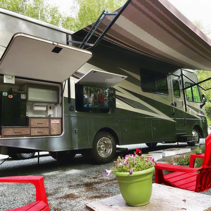 An RV rental setup at a campsite, featuring an outdoor kitchen, TV, and large awning. Red chairs ready for relaxation.. Winnebago Vista 2022
