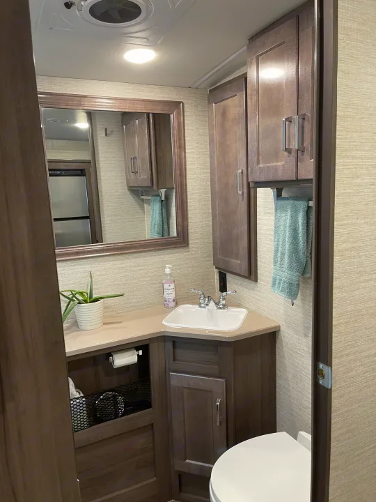 This compact RV rental bathroom features a corner sink, mirror, toilet, wood-toned cabinetry, and a ceiling vent.. Winnebago Vista 2022
