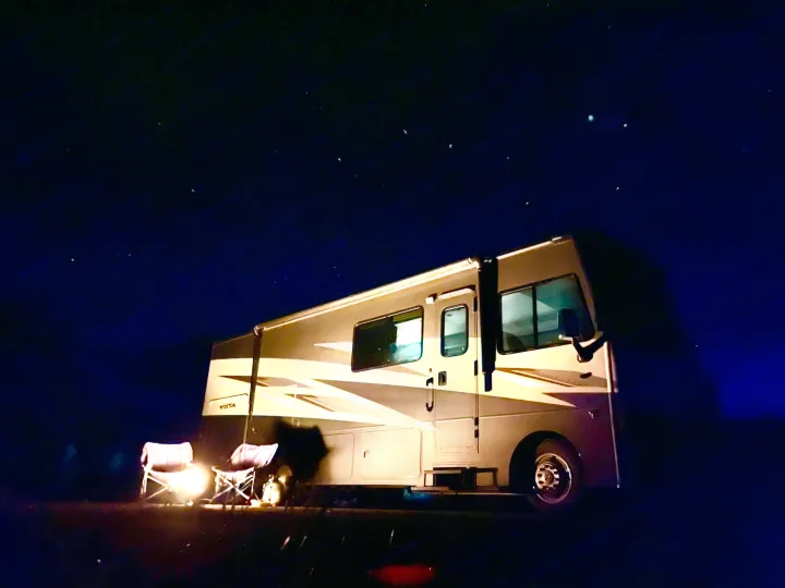 Stargazing with an RV rental. Cozy night camping under a vast, star-filled sky beside a large motorhome with lit chairs.. Winnebago Vista 2022