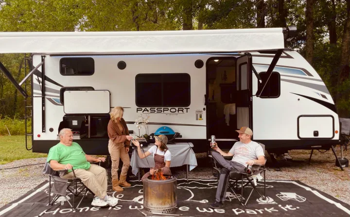 Four people gather around a spacious RV rental with an awning, fire pit, and grill setup for a cozy outdoor camping experience.. Keystone RV Passport SL 2021
