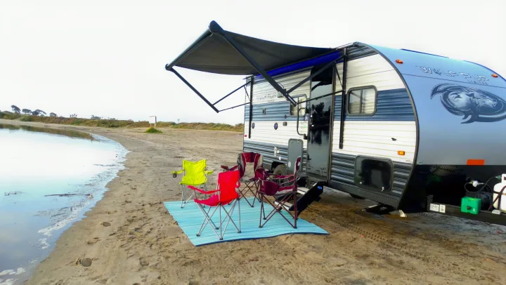 An RV rental is parked on a sandy shore next to calm water, with colorful chairs and a mat set up under its awning.. Forest River Cherokee Wolf Pup 2021