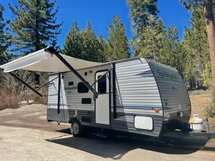 Springdale Mini travel trailer rv rental. A gray and white RV with an extended awning, parked at a sunny campsite amidst trees.. Keystone RV Springdale 2019