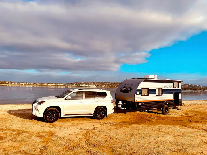 An RV rental, a Wolf Pup travel trailer, is towed by a white SUV on a sandy shore by a calm lake under a cloudy sky.. Forest River Cherokee Wolf Pup 2022