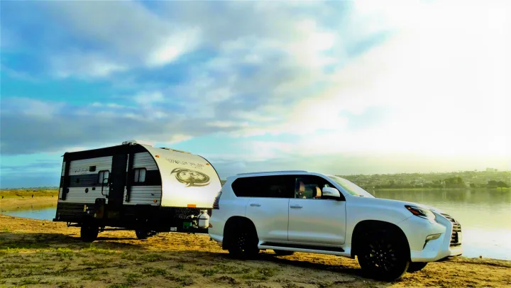 White SUV towing an RV rental trailer on a sandy shore by the water. Bright sky overhead for a perfect outdoor getaway.. Forest River Cherokee Wolf Pup 2022