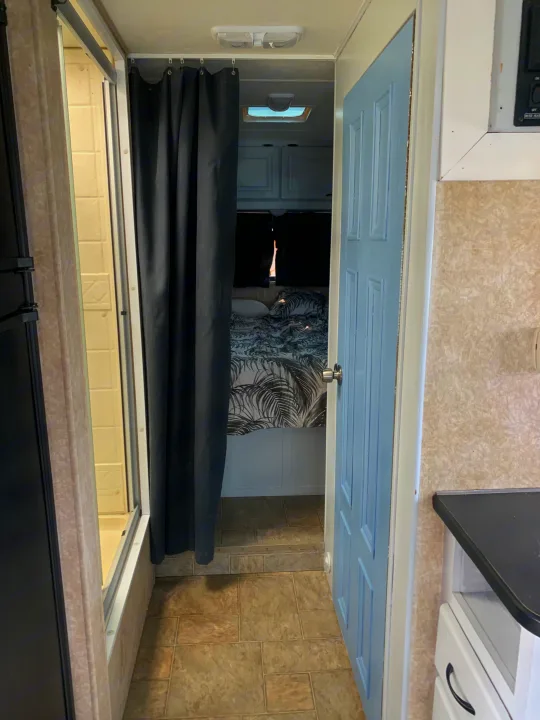 Experience this RV rental interior: An open light blue bedroom door, a bed with palm comforter, black privacy curtain, and kitchen counter.. Other Other 2008