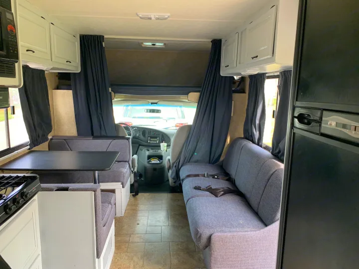Interior of an RV rental: a gray dinette, compact kitchen on left, and front cockpit. White cabinets & dark curtains.. Other Other 2008
