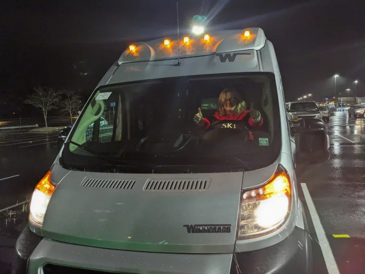 A happy woman gives two thumbs up from the driver's seat of a Winnebago RV rental van at night.. Winnebago Winnebago 2021