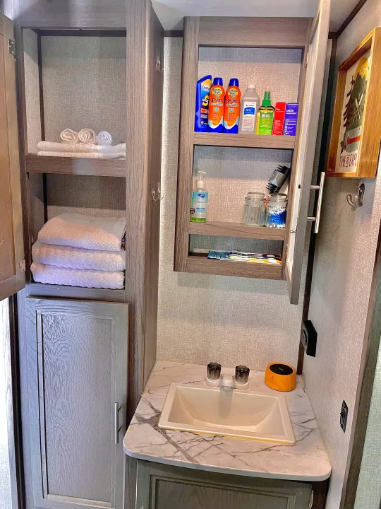 This RV rental bathroom features a sink, open medicine cabinet with toiletries, and a tall linen cabinet with white towels, maximizing compact storage.. Dutchmen Kodiak Ultra Lite 2021