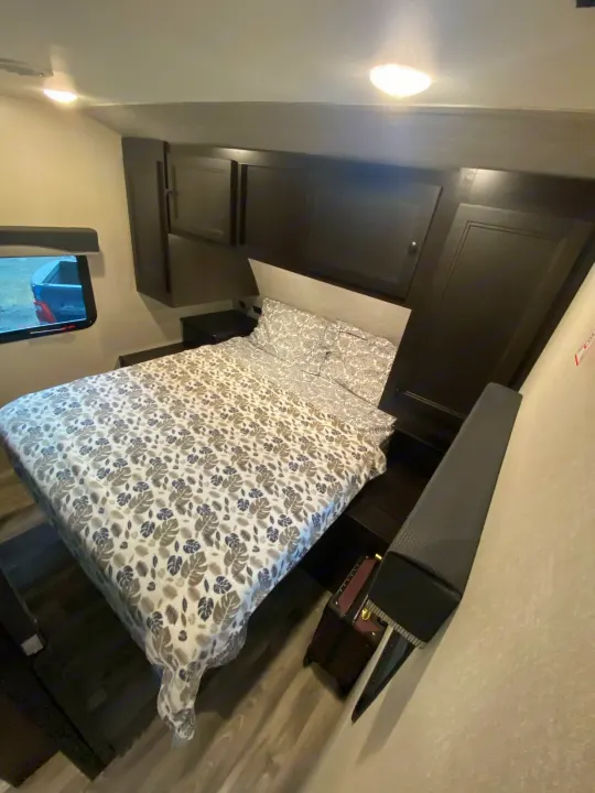 Cozy RV rental bedroom with leaf-patterned bedding, dark cabinets, and a window viewing a truck. Brightly lit interior.. Starcraft Super Lite 2022