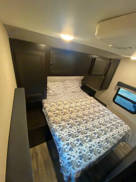 Cozy RV rental bedroom with leaf-patterned bedding, dark overhead cabinets, and a side window for compact comfort.. Starcraft Super Lite 2022