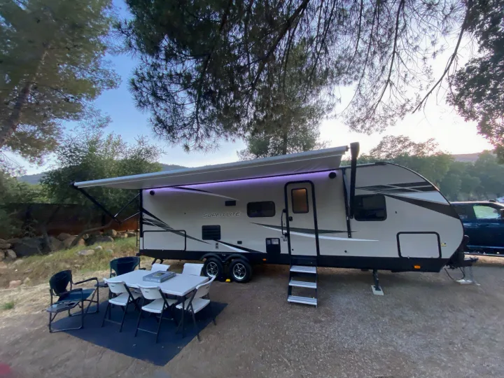 This RV rental is set up at a scenic campsite under trees, featuring an extended awning with purple lights, a dining table, and chairs.. Starcraft Super Lite 2022
