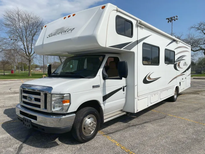 White Sunseeker Class C RV rental, a spacious motorhome perfect for travel, parked on a sunny lot near a sports field.. Forest River Sunseeker 2013
