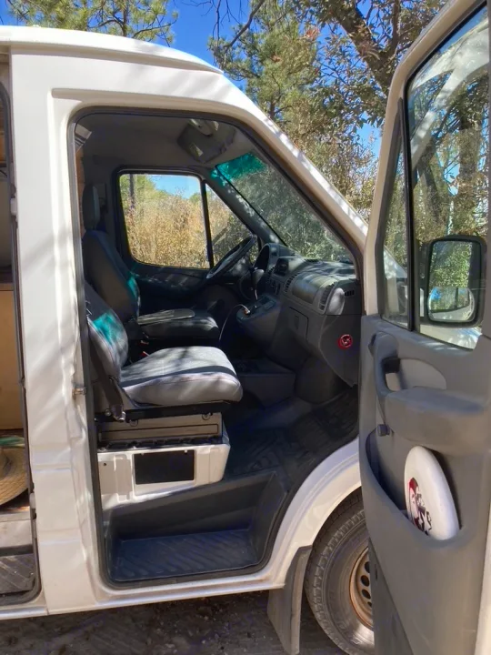 View inside an RV rental van, showcasing the driver's cabin with seats & steering wheel through the open door. Parked in a sunny, wooded area.. Dodge Sprinter 2003