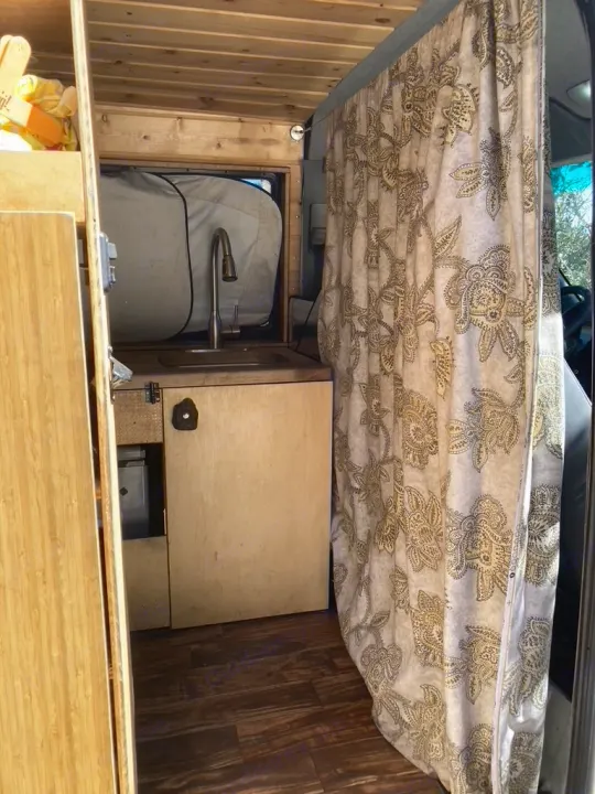 Inside an RV rental: A camper van's kitchen area with a sink, modern faucet, wood paneling, and a decorative privacy curtain.. Dodge Sprinter 2003