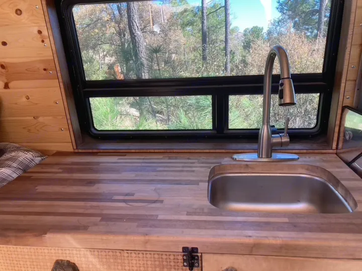 Inside an RV rental: a wooden kitchen counter with a stainless steel sink, pull-down faucet, and a large window view of the woods.. Dodge Sprinter 2003