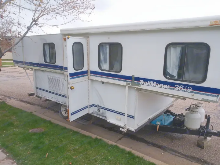 White TrailManor 2619 RV rental, partially expanded, parked on a street. Features include propane tanks.. K-Z Manufacturing Coyote 2002