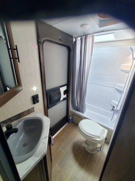 Compact RV rental bathroom: features a sink, toilet, tub with shower curtain, mirror, and an 'EXIT' sign.. Forest River Cherokee Grey Wolf 2021
