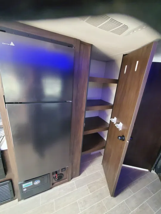 RV rental interior: Detailed view of a sleek refrigerator and open pantry with dark wood shelves in a compact kitchen.. Forest River Cherokee Grey Wolf 2021