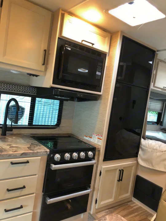 Modern RV rental kitchen with cream cabinets, black microwave, oven, large fridge, and sink. Stone-look counters.. Thor Four Winds 2022