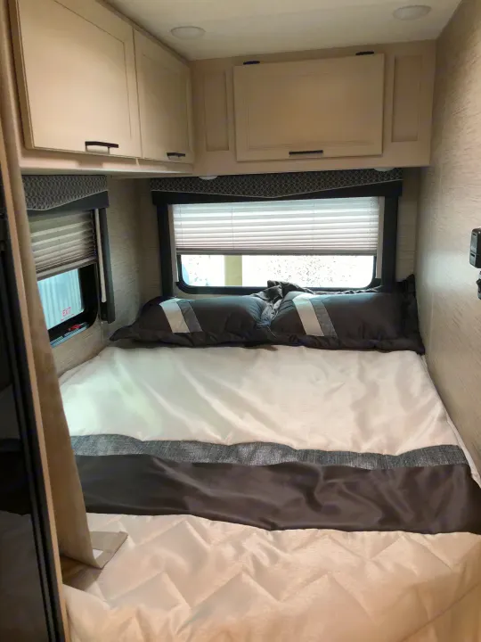 This compact RV rental bedroom features a comfy bed with white/grey bedding, overhead storage, and two large windows with blinds.. Thor Four Winds 2022