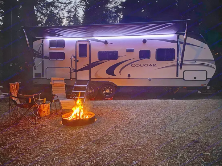 RV rental evening: A travel trailer with purple awning lights, a warm campfire, and chairs at night, perfect for camping.. Keystone Cougar Half-Ton 2022
