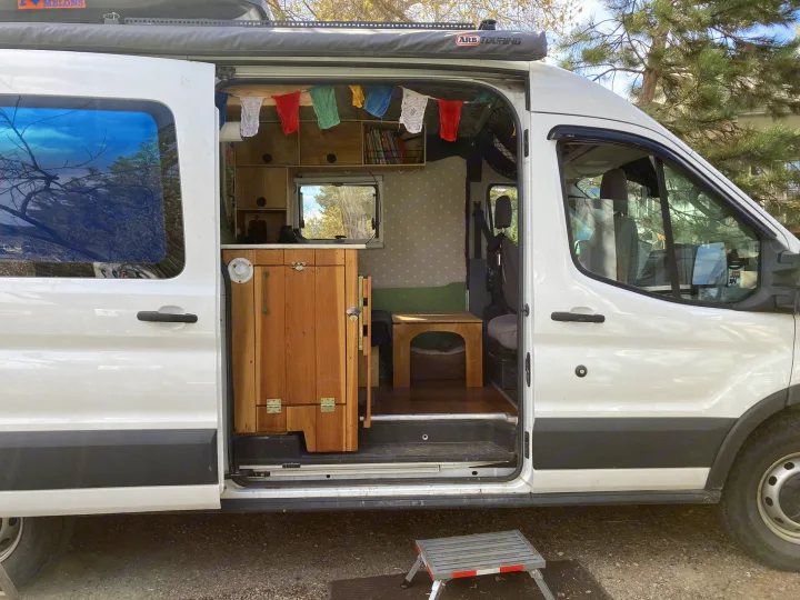 This white camper van, suitable for an rv rental, features an open side door revealing a custom wood interior & colorful prayer flags.. Ford Transit Custom 2015