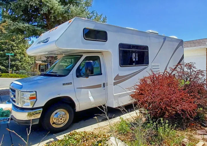 A white Class C motorhome (RV rental) parked on a sunny day, with trees & bushes nearby.. Thor Majestic 2015