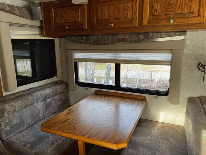 RV rental interior: A cozy dining area with a wooden table, patterned bench seating, and overhead cabinets. Windows show trees outside.. Allegro Allegro Bay 2004