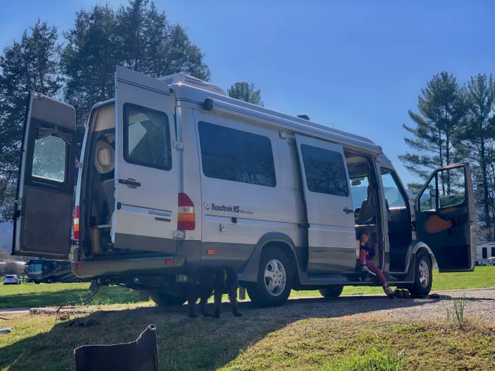 A silver Roadtrek RV rental is parked with open doors at a sunny campsite. A child is inside, two dogs stand near the back.. Roadtrek Roadtrek Class B 2007