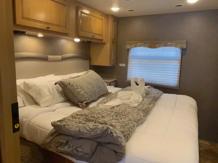 Inside an RV rental bedroom: a comfortable bed with swan towels, overhead cabinets, and a window with blinds.. Thor Windsport 2017