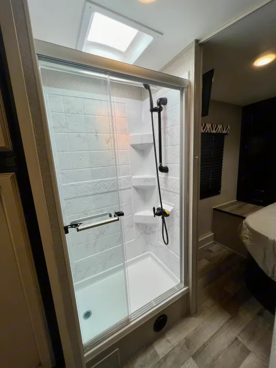 A modern, compact RV rental shower with white tiled-look walls, black fixtures, clear glass doors, and a skylight overhead.. Entegra Coach Odyssey 2021