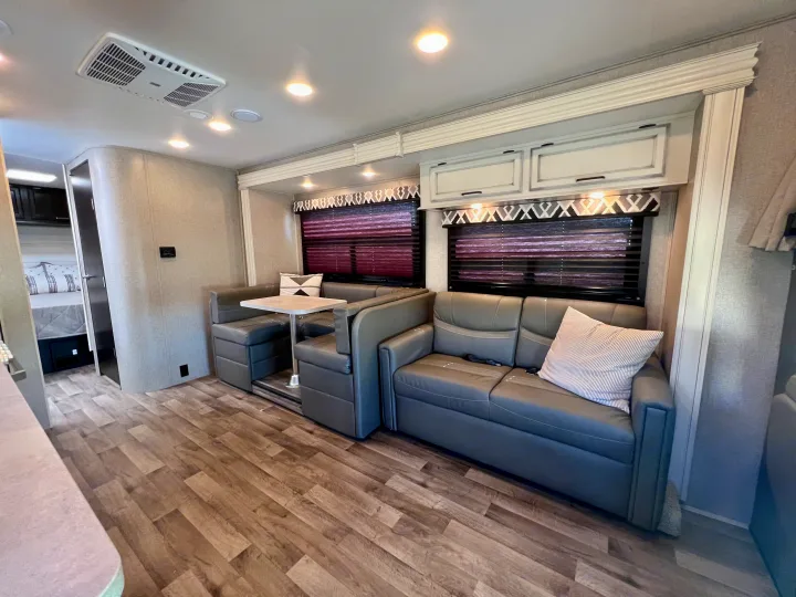 Modern RV rental interior with grey U-shaped dinette and sofa, light wood-look flooring, and overhead cabinets. Bedroom visible.. Entegra Coach Odyssey 2021