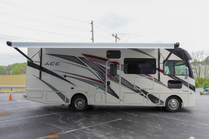 A white **RV rental** with an extended awning and modern graphics is parked on an asphalt lot.. Thor Motor Coach A.C.E 2022