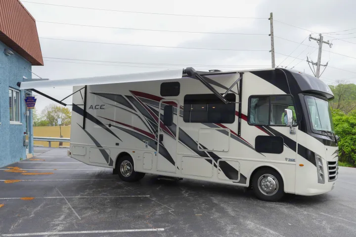 A white and black RV rental, possibly a Thor A.C.E., with an extended awning, parked in an outdoor lot.. Thor Motor Coach A.C.E 2022